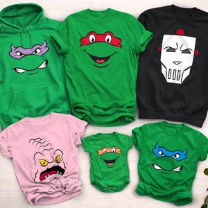 May include: A collection of green, pink, and black apparel featuring cartoon character faces. Items include a green hoodie, t-shirts, a baby onesie, and a black sweatshirt. The designs feature faces with colored masks and expressions.