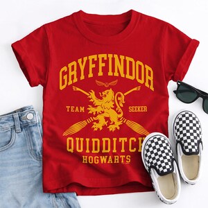 May include: A red t-shirt with the text "Gryffindor Quidditch Hogwarts" in yellow. The shirt features a lion emblem and two broomsticks. A pair of black and white checkered slip-on shoes and sunglasses are next to the shirt.