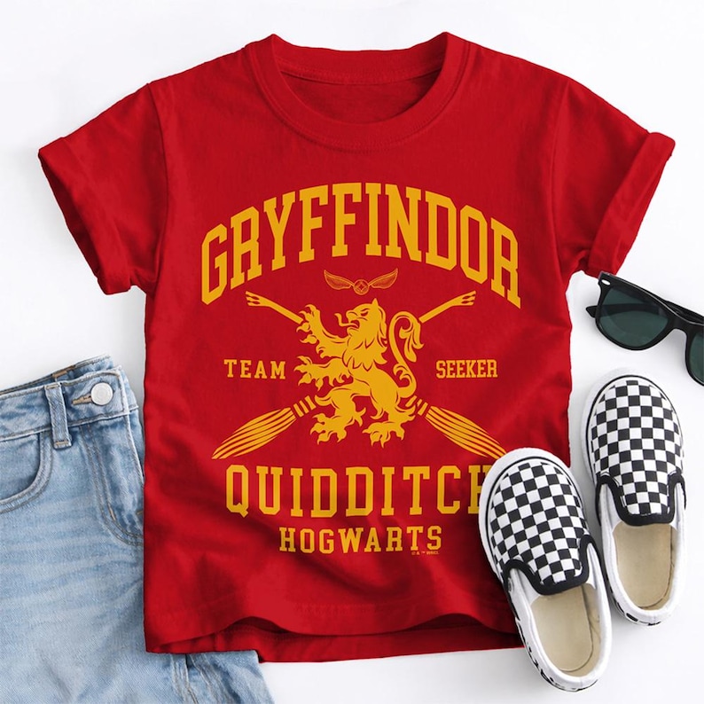 May include: A red t-shirt with the text "Gryffindor Quidditch Hogwarts" in yellow. The shirt features a lion emblem and two broomsticks. A pair of black and white checkered slip-on shoes and sunglasses are next to the shirt.