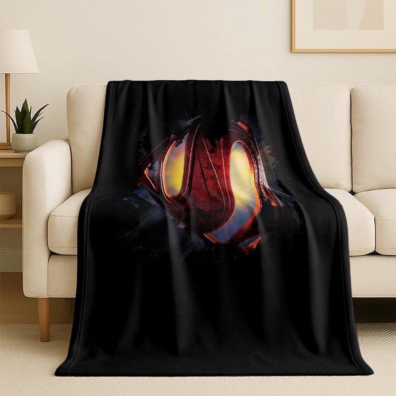 Puede incluir: Manta de forro polar negra con un gran logotipo de Superman en rojo, amarillo y azul. El logotipo se encuentra sobre un fondo oscuro y texturizado. La manta est&aacute; sobre un sof&aacute; beige, creando un contraste llamativo.