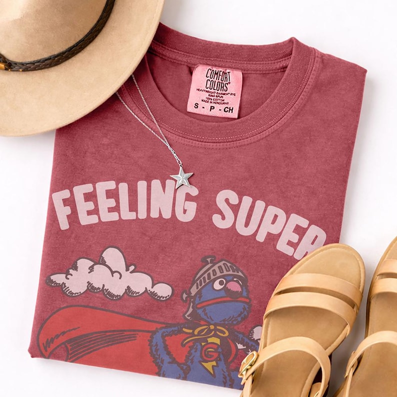 May include: A maroon t-shirt with the words "FEELING SUPER" in light pink, featuring a cartoon character in a superhero costume. A silver star necklace, a tan hat, and tan sandals are also in the image.