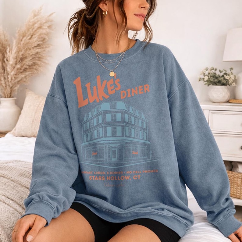 Op de afbeelding: Een blauwe crewneck sweatshirt met de tekst "Luke's Diner" in oranje, boven een gedetailleerde illustratie van een diner. Daaronder staat de tekst "BREAKFAST LUNCH & DINNER NO CELL PHONES STARS HOLLOW, CT."