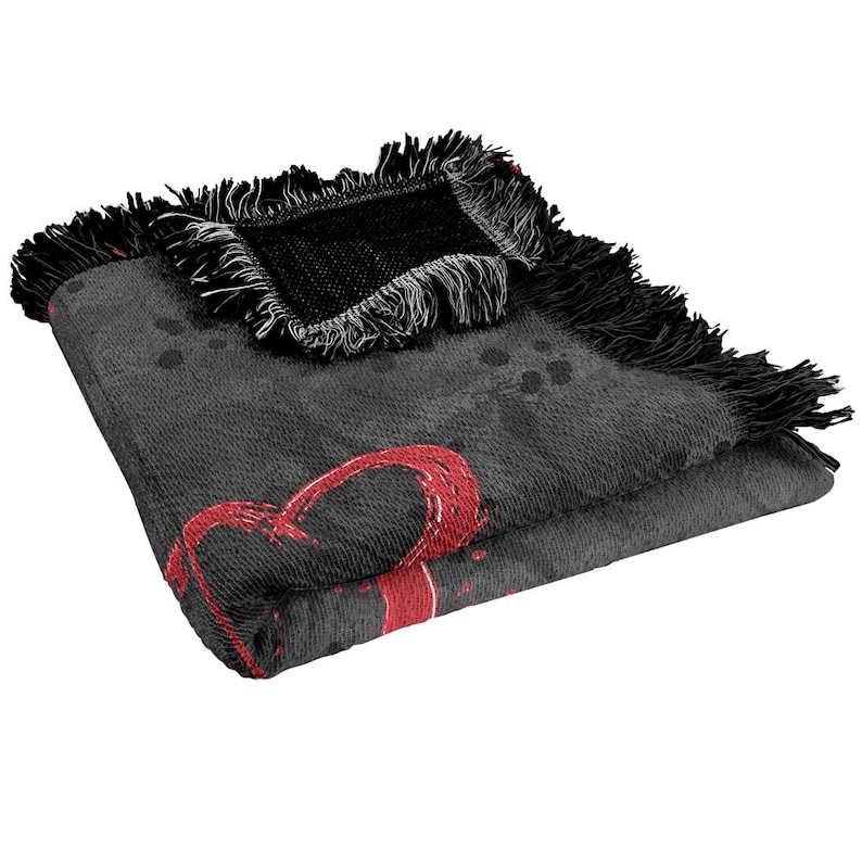 May include: Folded gray throw blanket with a large red heart design. The blanket has black fringe trim and a black woven patch. The heart design appears to be hand-drawn. The blanket is folded over on itself.