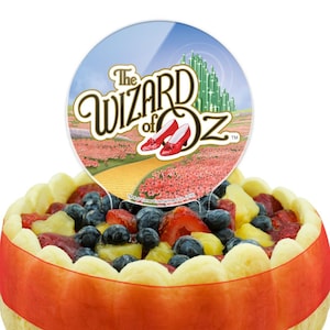 May include: A fruit tart with a red ribbon and a decorative topper featuring the words "The Wizard of Oz", ruby slippers, and a yellow brick road. The tart is filled with blueberries, strawberries, and pineapple.