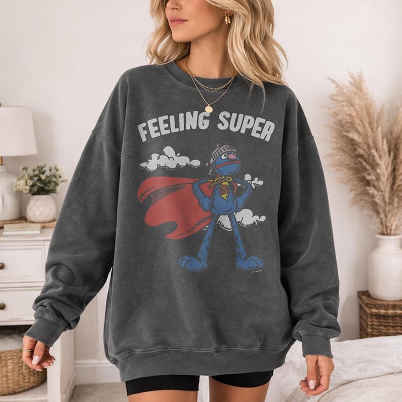 May include: Dark gray sweatshirt with the words "FEELING SUPER" above a cartoon superhero character. The superhero is blue with a red cape and a helmet. The sweatshirt is worn by a person.