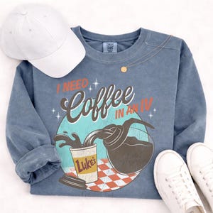 May include: A blue-grey sweatshirt with the phrase "I NEED Coffee IN AN IV" printed on the front. The graphic depicts coffee pouring into a cup from a carafe. A white baseball cap and white sneakers are also in the image.
