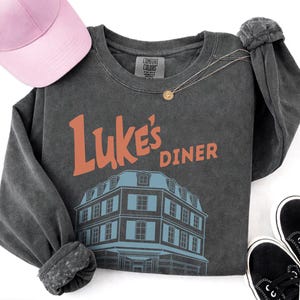 Op de afbeelding: Donkergrijs shirt met lange mouwen met de oranje tekst "Luke's Diner" en een blauwe illustratie van een restaurant. Een roze baseballpet, een gouden ketting en zwarte sneakers zijn ook zichtbaar.