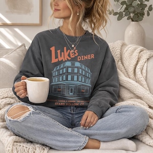 Op de afbeelding: Een donkergrijs sweatshirt met de tekst "Luke's Diner" in oranje en een blauwe illustratie van een diner. Het sweatshirt wordt gecombineerd met een lichtblauwe jeans en een witte mok. De tekst "BREAKFAST, LUNCH, & DINNER - NO CELL PHONE STARS HOLLOW, CT" is ook zichtbaar.