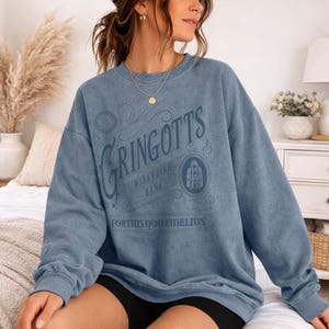 May include: A blue sweatshirt with the text "Gringotts Wizarding Bank" in a vintage style. The design includes an illustration of a building and the phrase "Fortius Quo Fidelius". The sweatshirt is a solid blue color.