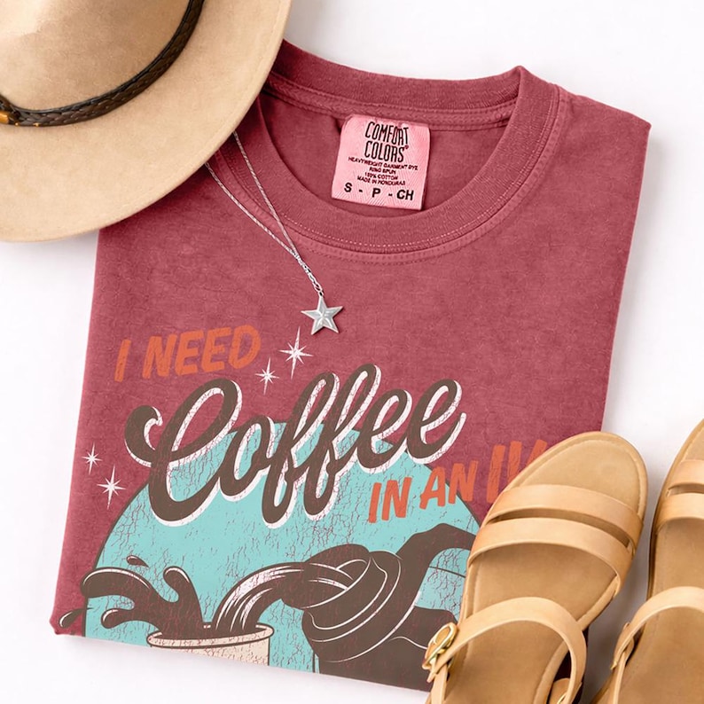 May include: A maroon t-shirt with the text "I NEED Coffee IN AN IV" and a coffee-themed graphic. A silver star pendant necklace, a beige hat, and tan sandals are also in the image.