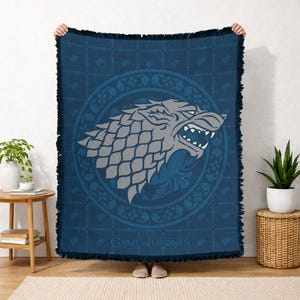 May include: A dark blue throw blanket featuring a gray direwolf head within a circular design. The blanket has a fringed edge and the words "GAME OF THRONES" are printed at the bottom. The direwolf is the emblem of House Stark.