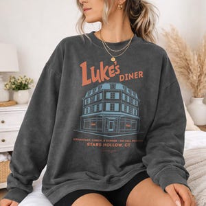 Op de afbeelding: Donkergrijze sweatshirt met de oranje tekst "Luke's Diner" en een illustratie van een diner. De tekst eronder luidt "Breakfast, Lunch, & Dinner - No Cell Phones, Stars Hollow, CT". De sweatshirt heeft een casual stijl.
