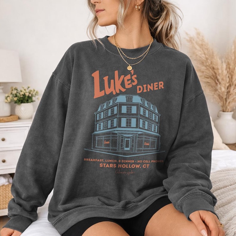 Op de afbeelding: Donkergrijze sweatshirt met de oranje tekst "Luke's Diner" en een illustratie van een diner. De tekst eronder luidt "Breakfast, Lunch, & Dinner - No Cell Phones, Stars Hollow, CT". De sweatshirt heeft een casual stijl.