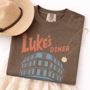 Op de afbeelding: Een bruin t-shirt met de tekst "Luke's Diner" in oranje en een blauw-witte gebouwafbeelding. Een beige hoed met een bruine band en een madeliefjesketting zijn ook zichtbaar.