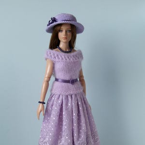 Könnte beinhalten: Eine Puppe in einem lavendelfarbenen Outfit, bestehend aus einem Hut, einem schulterfreien Oberteil und einem plissierten Rock mit floralem Spitzenbesatz. Das Outfit ist mit einem passenden Gürtel, einer Halskette und einem Armband ausgestattet. Die Puppe hat braunes Haar.