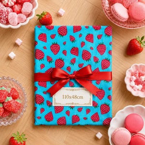 Peut inclure: Une pièce de tissu rectangulaire sur fond bleu vif avec un motif répétitif de fraises rouges. Un ruban de satin rouge est noué en nœud au centre, retenant une étiquette rectangulaire blanche avec le texte imprimé 110x48cm.