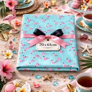 Peut inclure: Un tissu turquoise plié, orné d'un motif de flamants roses, noué avec un ruban rose et un nœud noir. L'étiquette indique "70 x 61cm". La scène comprend des tasses à thé, des fleurs et des éléments décoratifs, sur le thème tropical.