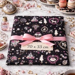Peut inclure: Un tissu noir, mesurant 70 x 33 cm, est enveloppé d'un ruban et d'un nœud roses. Le tissu est orné d'illustrations roses et blanches sur le thème des contes de fées. La scène comprend des tasses à thé, des pâtisseries et des flacons de parfum.