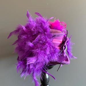 Puede incluir: Un sombrero morado vibrante con una profusi&oacute;n de plumas y un acento rosa. El sombrero presenta un ala estructurada y est&aacute; adornado con adornos decorativos y una flor de tela. El sombrero se exhibe sobre un soporte negro.
