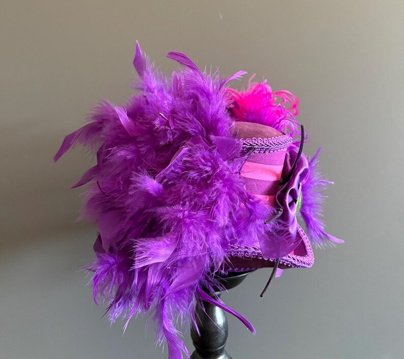 Puede incluir: Un sombrero morado vibrante con una profusi&oacute;n de plumas y un acento rosa. El sombrero presenta un ala estructurada y est&aacute; adornado con adornos decorativos y una flor de tela. El sombrero se exhibe sobre un soporte negro.