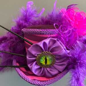 Puede incluir: Un sombrero de copa morado y rosa con un broche en forma de ojo. El sombrero presenta una flor de sat&eacute;n morada, plumas y adornos. El broche tiene un dise&ntilde;o de ojo verde y amarillo.