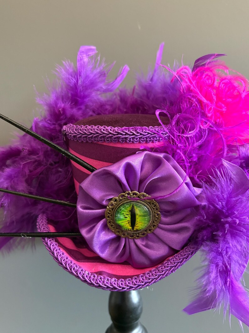 Puede incluir: Un sombrero de copa morado y rosa con un broche en forma de ojo. El sombrero presenta una flor de sat&eacute;n morada, plumas y adornos. El broche tiene un dise&ntilde;o de ojo verde y amarillo.