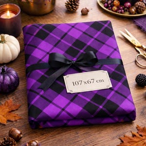 Peut inclure: Un tissu plié à carreaux violets et noirs, noué avec un ruban noir. Une étiquette sur le tissu indique "107 x 67 cm". La scène comprend des décorations d'automne, comme des bougies, des citrouilles et des feuilles.