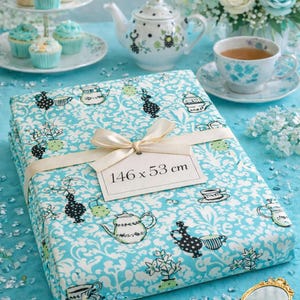Peut inclure: Un tissu turquoise orné de motifs de théières et de tasses, noué avec un ruban crème et une étiquette indiquant "146 x 53 cm". La scène comprend une théière, une tasse, des cupcakes et des fleurs, tous dans des tons de bleu et de blanc, sur le thème du tea party.
