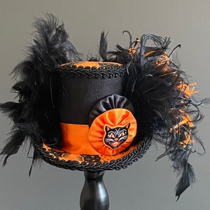 May include: A black top hat with orange and black trim and a black cat face appliqu&eacute;. The hat is adorned with black feathers.