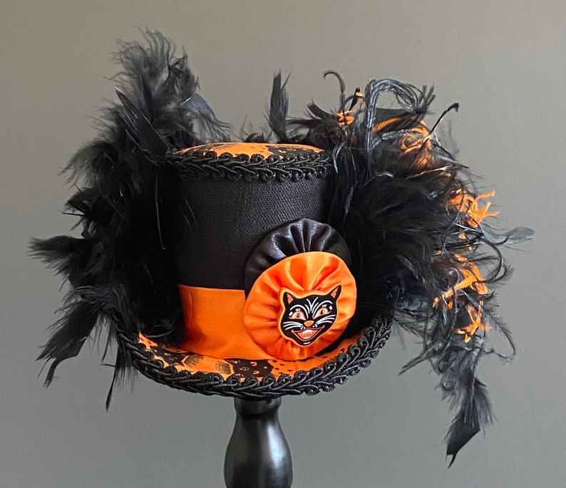 May include: A black top hat with orange and black trim and a black cat face appliqu&eacute;. The hat is adorned with black feathers.