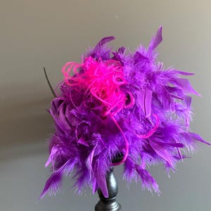 Puede incluir: Un fascinador de plumas moradas vibrantes con un acento rosa brillante. El tocado est&aacute; compuesto por plumas en capas, creando una apariencia voluminosa y texturizada. El dise&ntilde;o est&aacute; montado sobre una base oscura, adecuado para ocasiones especiales.