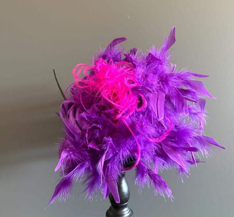 Puede incluir: Un fascinador de plumas moradas vibrantes con un acento rosa brillante. El tocado est&aacute; compuesto por plumas en capas, creando una apariencia voluminosa y texturizada. El dise&ntilde;o est&aacute; montado sobre una base oscura, adecuado para ocasiones especiales.