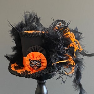 May include: A black top hat with an orange and black ribbon and a black cat face appliqu&eacute;. The hat is decorated with black and orange feathers.