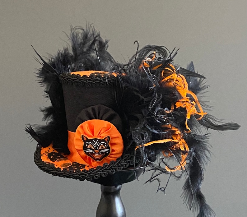 May include: A black top hat with an orange and black ribbon and a black cat face appliqu&eacute;. The hat is decorated with black and orange feathers.