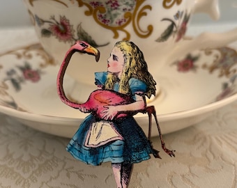 May include: A whimsical wooden brooch featuring a depiction of Alice in Wonderland holding a pink flamingo. The brooch is set against a backdrop of a floral teacup and saucer, evoking a sense of fantasy and storytelling.