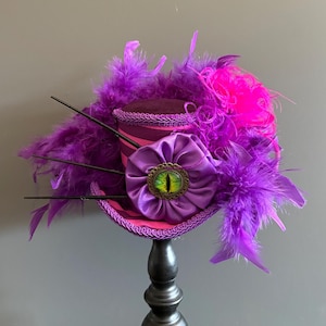 Puede incluir: Un sombrero de copa morado y rosa con una gran flor de sat&eacute;n morado y un adorno en forma de ojo. El sombrero est&aacute; adornado con plumas moradas y rosas y detalles en negro. El sombrero se exhibe sobre un soporte negro.