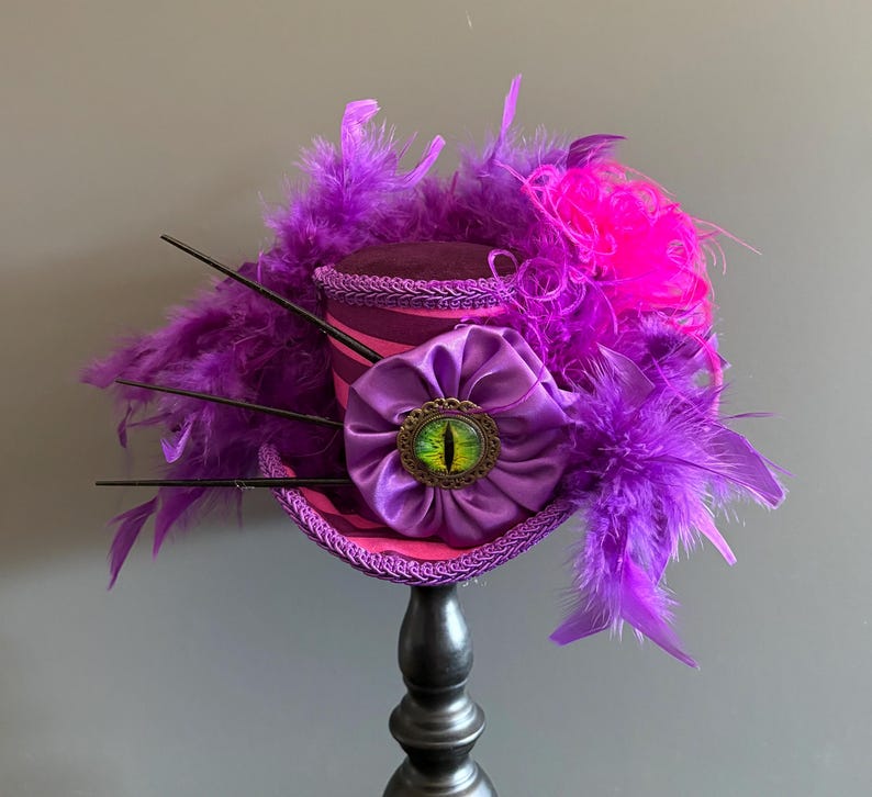 Puede incluir: Un sombrero de copa morado y rosa con una gran flor de sat&eacute;n morado y un adorno en forma de ojo. El sombrero est&aacute; adornado con plumas moradas y rosas y detalles en negro. El sombrero se exhibe sobre un soporte negro.
