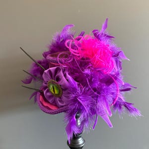 Puede incluir: Tocado decorativo morado y rosa, con una flor central con un detalle verde en forma de ojo. El dise&ntilde;o incorpora plumas moradas y detalles rosas, creando una est&eacute;tica teatral y caprichosa.