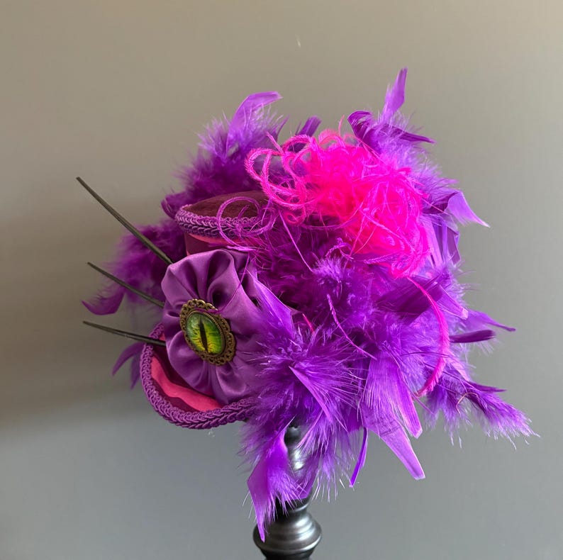 Puede incluir: Tocado decorativo morado y rosa, con una flor central con un detalle verde en forma de ojo. El dise&ntilde;o incorpora plumas moradas y detalles rosas, creando una est&eacute;tica teatral y caprichosa.