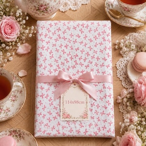 Peut inclure: Un tissu blanc orné de motifs de nœuds roses, noué avec un ruban rose et une étiquette. L'étiquette indique "114x98cm". L'image est entourée de tasses à thé, de roses et de macarons, créant une esthétique vintage.