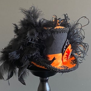 May include: A black top hat with orange and black trim and a black feather boa. The hat has a black and white cat silhouette and a black and orange bow.