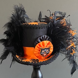 May include: A black top hat with orange and black trim and a black cat face appliqu&eacute;. The hat is decorated with black feathers.
