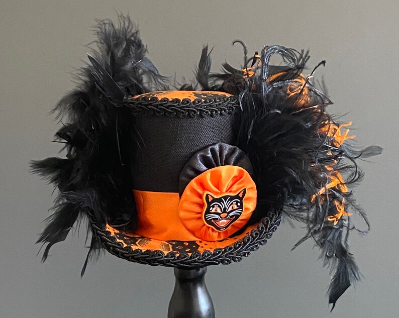 May include: A black top hat with orange and black trim and a black cat face appliqu&eacute;. The hat is decorated with black feathers.
