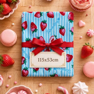 Peut inclure: Un morceau de tissu rectangulaire avec un motif à rayures verticales bleu clair et blanc, des illustrations de fraises rouges et le mot Strawberry imprimé. Il est noué avec un ruban rouge et une étiquette affichant le texte 115x53cm.