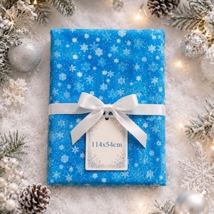 Peut inclure: Un cadeau emballé dans du papier bleu à motifs flocons de neige, noué avec un ruban et un nœud blancs. Une étiquette indique les dimensions 114x54cm. La scène festive comprend des décorations de Noël, des pommes de pin et des guirlandes lumineuses.