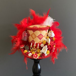 Peut inclure: Un chapeau haut de forme décoratif avec un motif de losanges rouges et dorés, orné de plumes rouges, de roses miniatures et de figurines de personnages. Le chapeau comprend une petite cloche en verre avec une rose à l'intérieur et une figurine d'horloge.