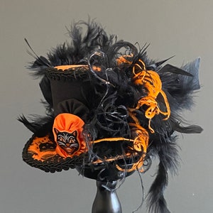 May include: A black and orange top hat with black feathers and a black cat face on the front. The hat is decorated with black lace and orange ribbon.