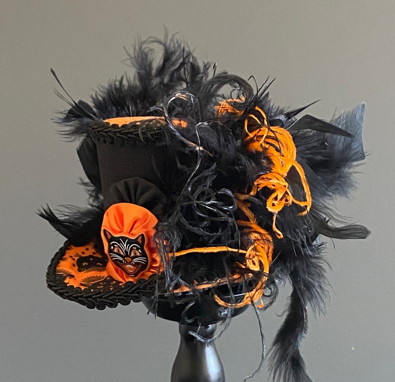 May include: A black and orange top hat with black feathers and a black cat face on the front. The hat is decorated with black lace and orange ribbon.
