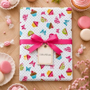 Peut inclure: Une pièce de tissu rectangulaire à fond blanc avec un motif à pois gris, ornée de cupcakes, de tasses à thé et de papillons colorés. Un ruban rose vif est noué au centre avec une étiquette indiquant les dimensions 110 x 92 cm.