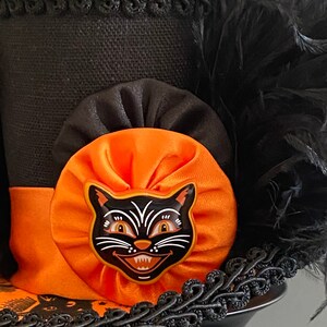 May include: A black top hat with an orange and black bow and a black cat face appliqu&eacute;. The hat is decorated with black feathers and black trim.